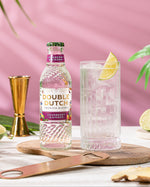 Bottle of Double Dutch CRANBERRY & GINGER TONIC WATER with a glass of gin and tonic on a table with a pink background