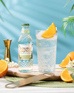 Bottle of Double Dutch ELDERFLOWER TONIC WATER (24 x 200ml) with a glass of gin and tonic garnished with orange slices on a table with oranges and plants.