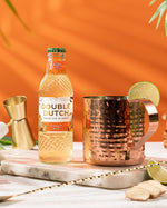 Double Dutch GINGER ALE with copper mugs and lime wedges on a wooden surface against an orange background