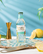 Bottle of Double Dutch REFRESHING LEMONADE with lemons and a jigger on a table against a blue background