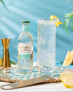 Bottle of Double Dutch REFRESHING LEMONADE with a glass of gin and tonic on a table with lemons and a blue background