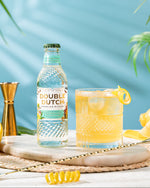 Bottle of Double Dutch REFRESHING LEMONADE with a cocktail garnished with lemons on a table with a blue background.